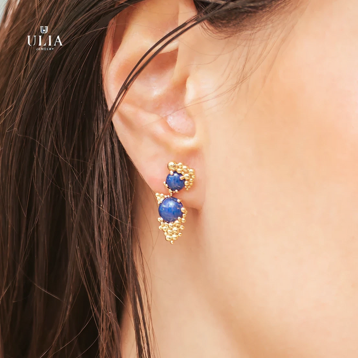 Gold earrings with blue gemstones worn by a person, brand 'ULIA' visible.